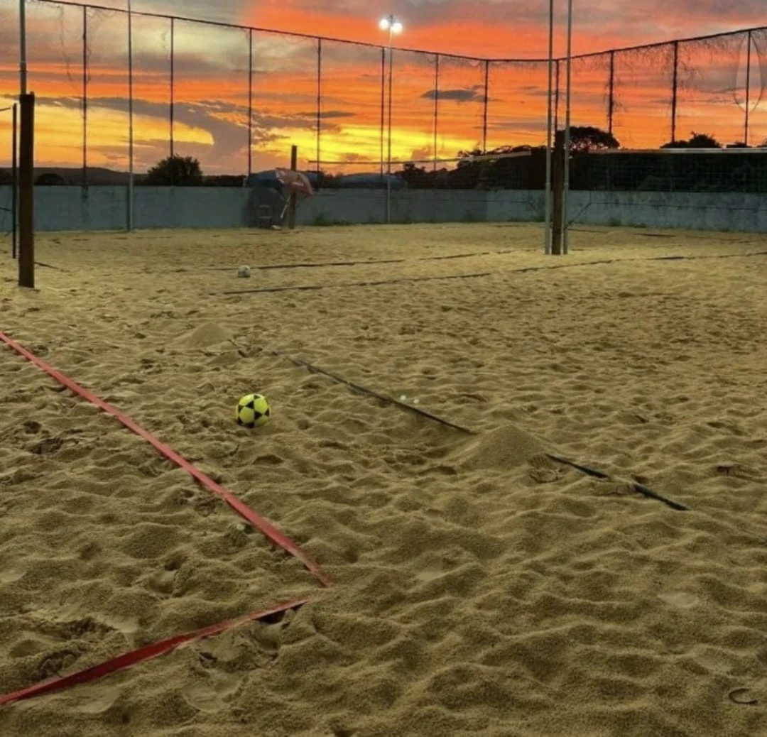 Quadras de areia da Arena CHT iluminadas ao pôr do sol, com bola de futevôlei em primeiro plano